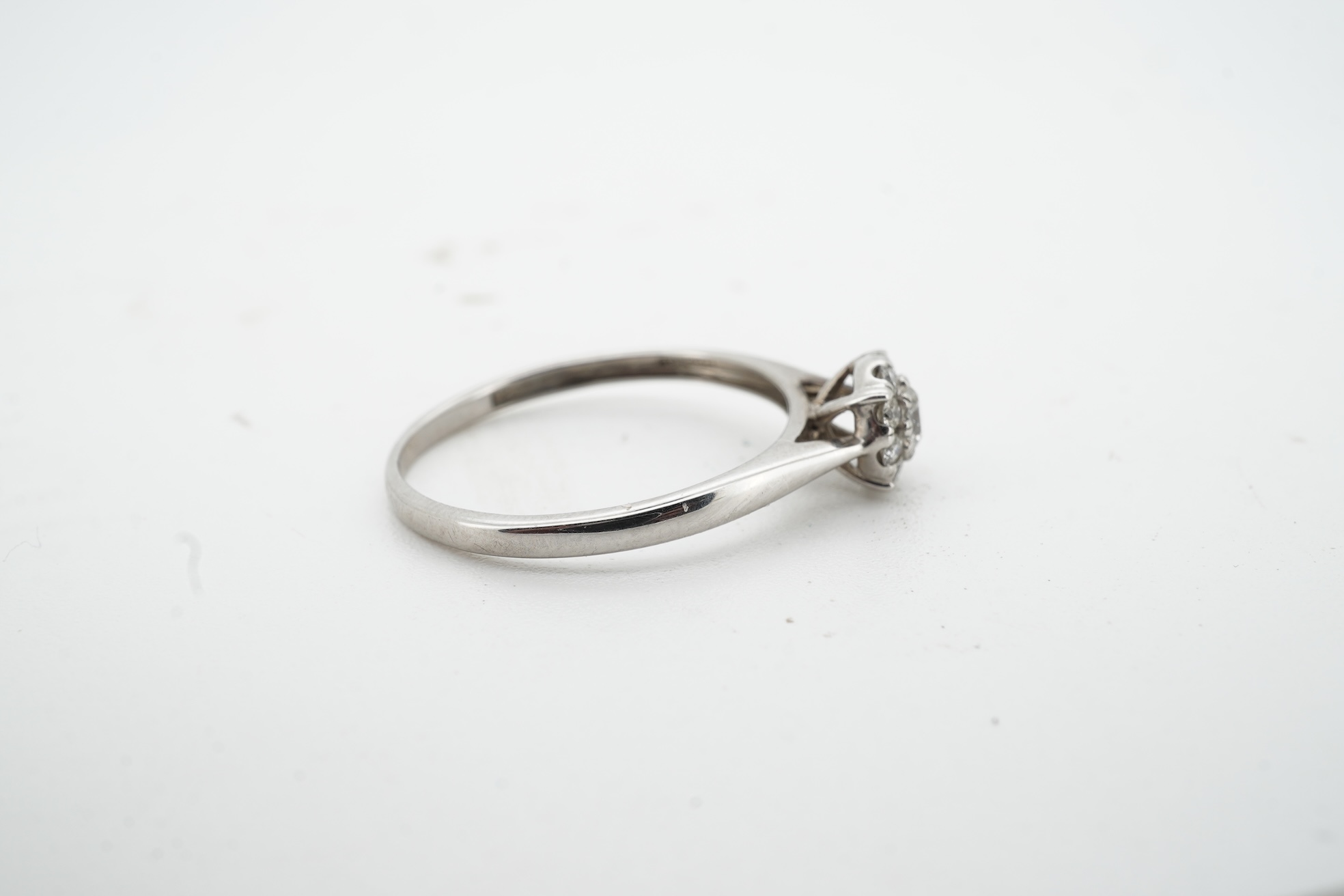 A diamond cluster ring, claw-set with a brilliant-cut diamond weighing approximately 0.10 carats, within a border of smaller brilliant-cut diamonds totalling approximately 0.09 carats, mounted in unmarked white metal, si